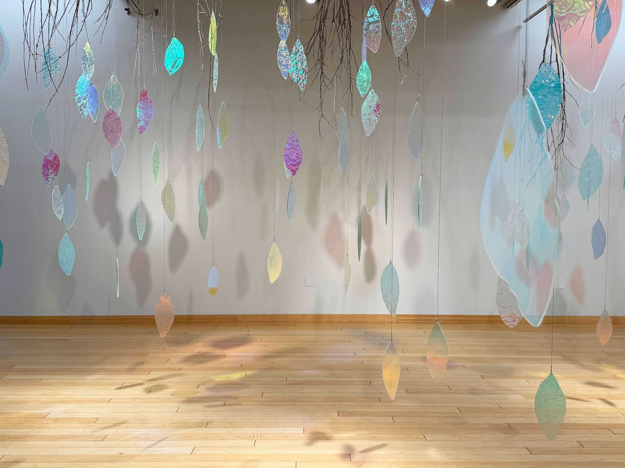 Dichroic glass leaf-shaped forms hanging from strings and branches in a gallery installation