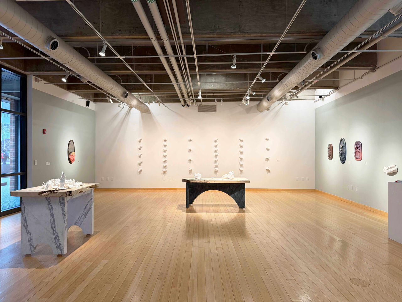 Wide view of a gallery space with sculpted wall pieces and two large handmade tables displaying white plaster hand sculptures