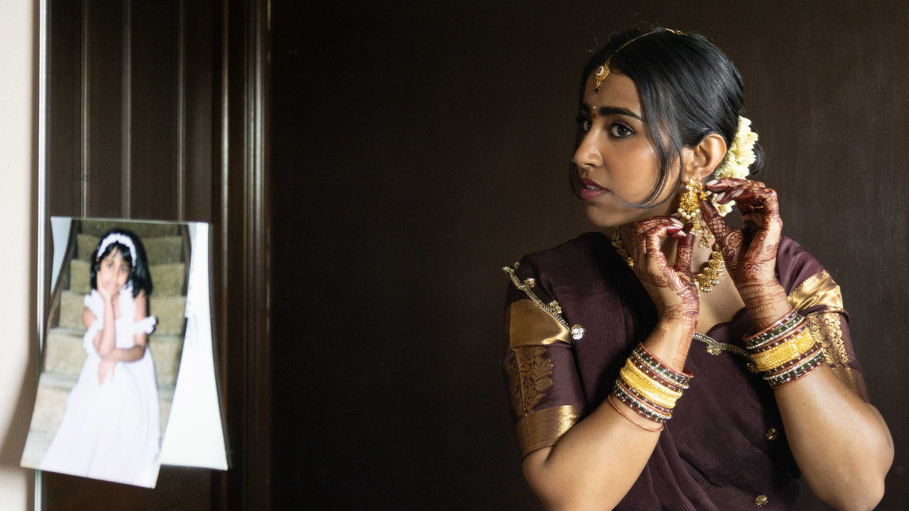 Color portrait photograph of an Indian woman putting on earrings next to a photograph of a young child, titled "Younger Self" by Vidushi Shrivastava, 2026; photograph in UC DAAP Galleries' Reed Gallery Exhibit Long Exposure