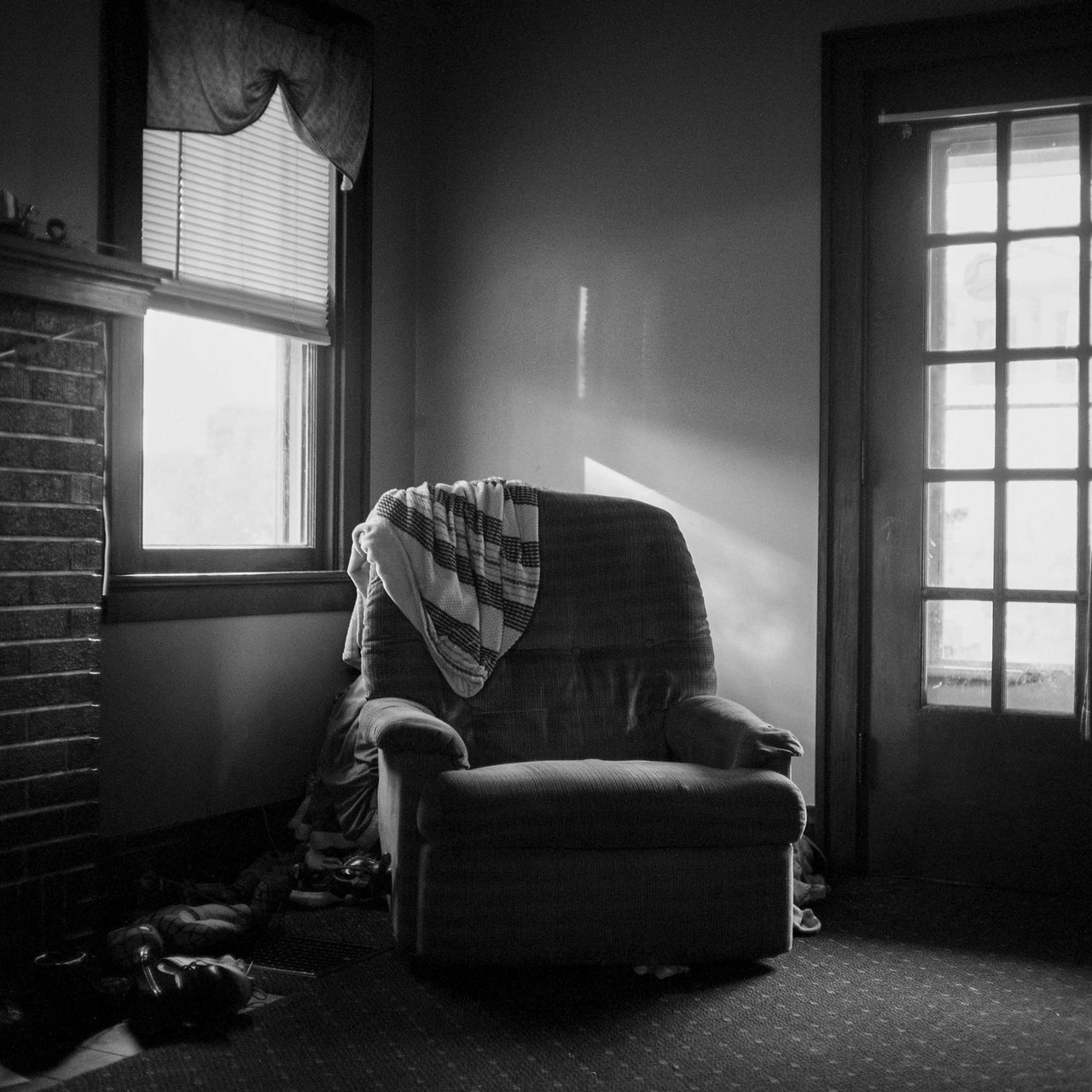 Black-and-white photograph of empty sitting chair with trow-blanket next to a window titled "Mother's Chair" by Grant Lawler, 2021; photograph in UC DAAP Galleries' Reed Gallery Exhibit Long Exposure