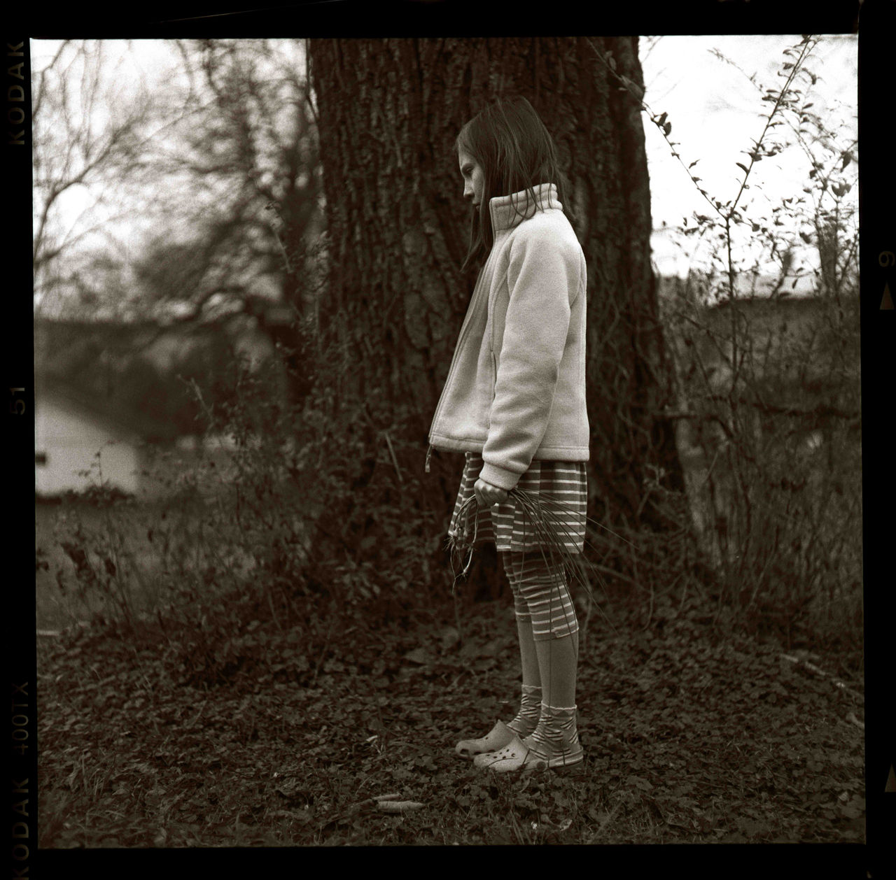 Sepia-toned side-profile photograph of a young child standing beside a large tree in a wooded area, wearing a light fleece jacket, striped shorts, leggings, and Crocs-style shoes while holding a bundle of long grass or thin branches in one hand. Dense vegetation and tree trunks surround the child. Title "Onions" by Robert Hein, 2013; photograph in UC DAAP Galleries' Reed Gallery Exhibit Long Exposure