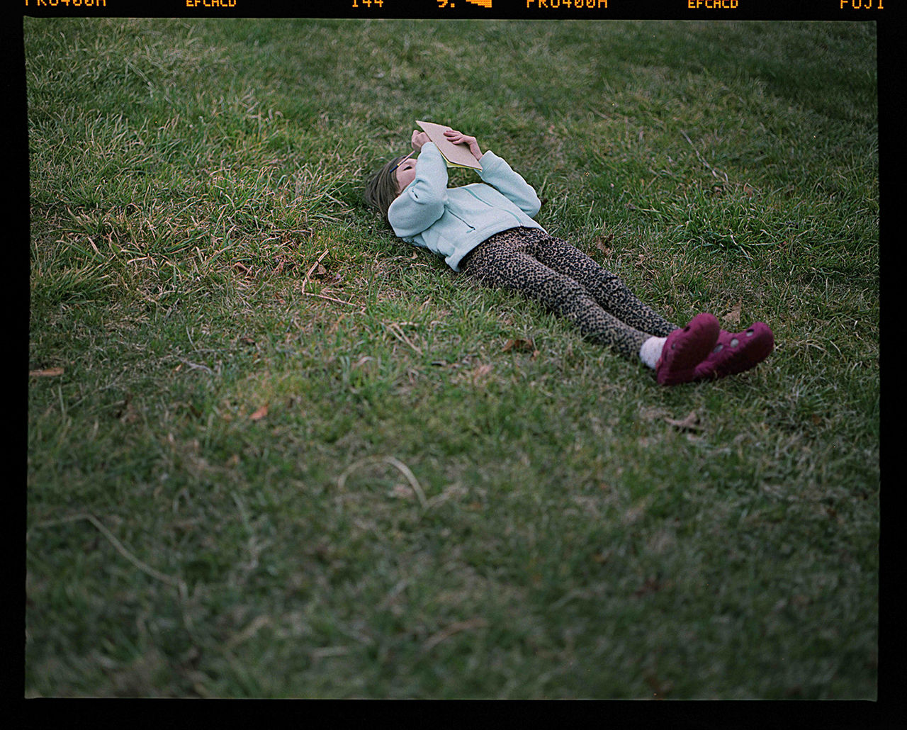 Color photograph of a young child lying on their back in a grassy field and holding an open book above their face. The child wears a light blue jacket, leopard-print pants, white socks, and bright pink Crocs-style shoes, with the surrounding grass filling most of the frame. Titled "Notes" by Robert Hein, 2016; photograph in UC DAAP Galleries' Reed Gallery Exhibit Long Exposure