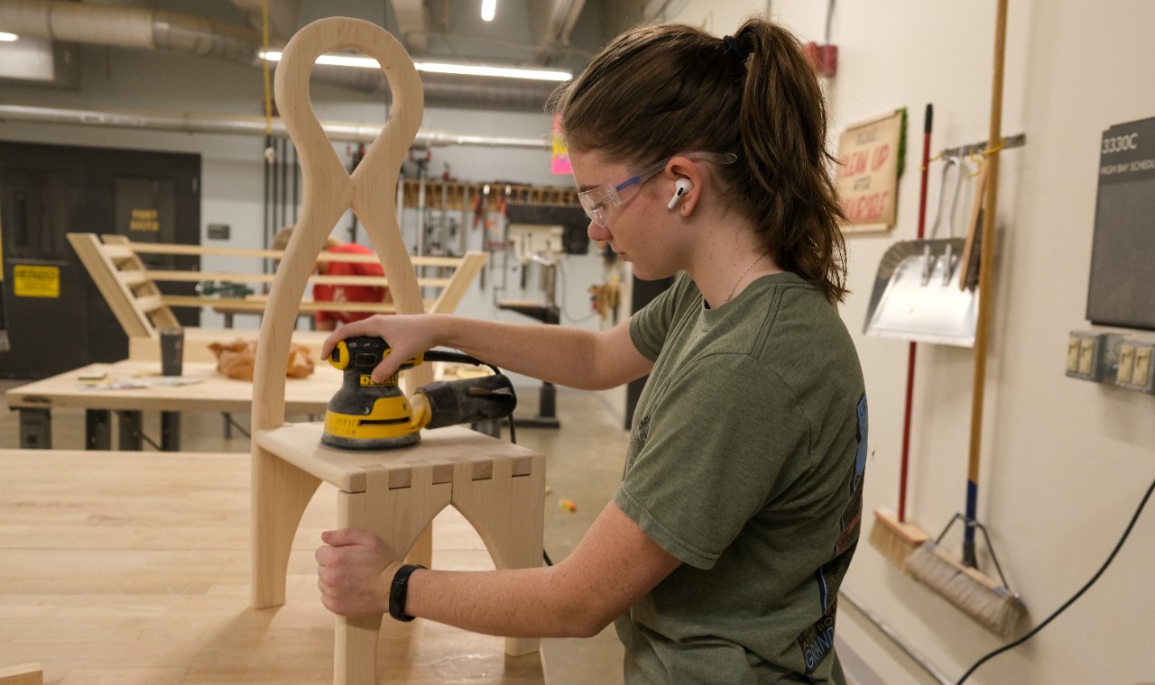 A female student wearing a grey blazer and hair in a bun stands next to a male student with curly hair, a beard and wearing glasses and a sweater as they both look at a big white board with lots of multi-color post-it notes on it